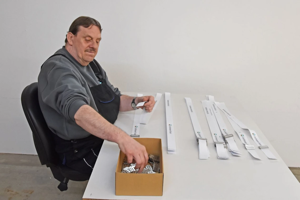 Ein Mann in grauem Pullover und Arbeitshose sitzt an einem Tisch und schiebt Metallteile aus einer braunen Box auf wei&szlig;e Papier- oder Stoffstreifen.