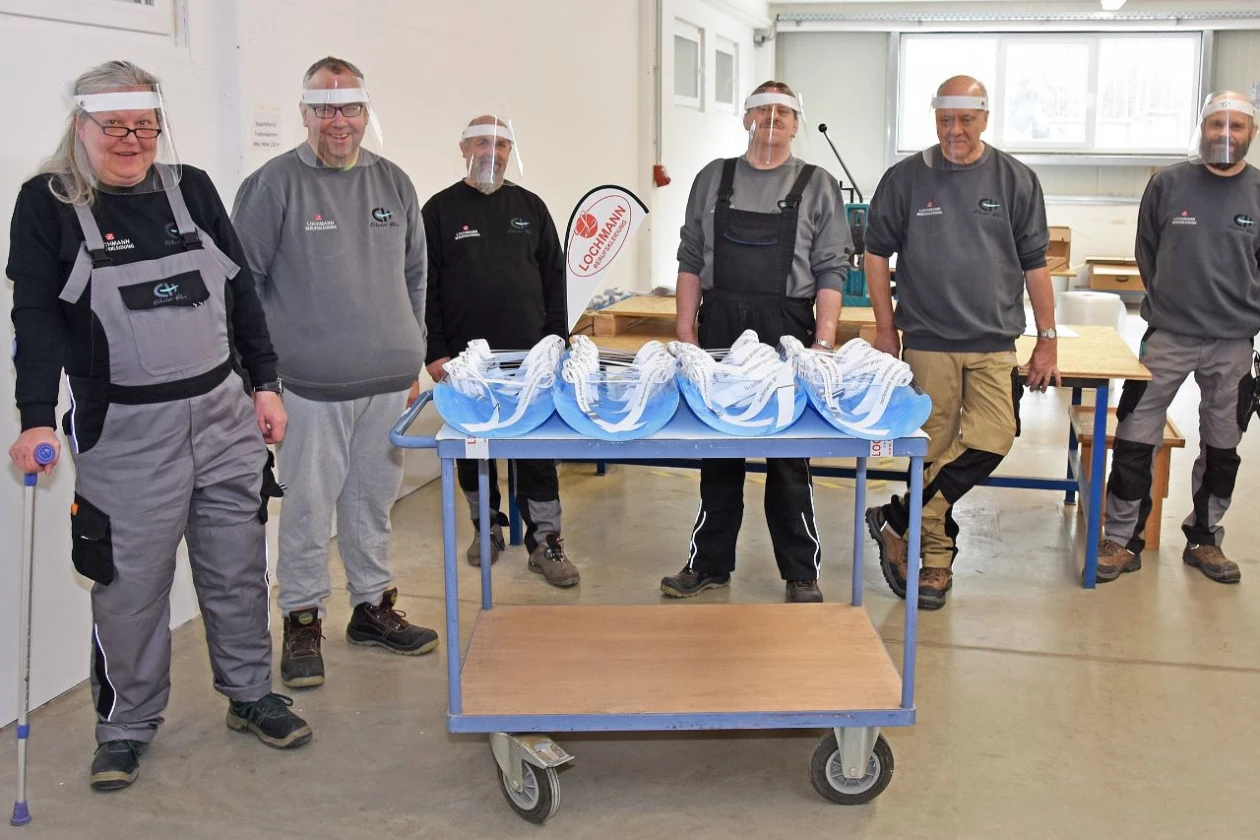 Sechs Personen in Arbeitskleidung mit Gesichtsvisieren stehen hinter einem Transportwagen mit blauen Gesichtsvisieren. Im Hintergrund sind Holztische zu sehen.