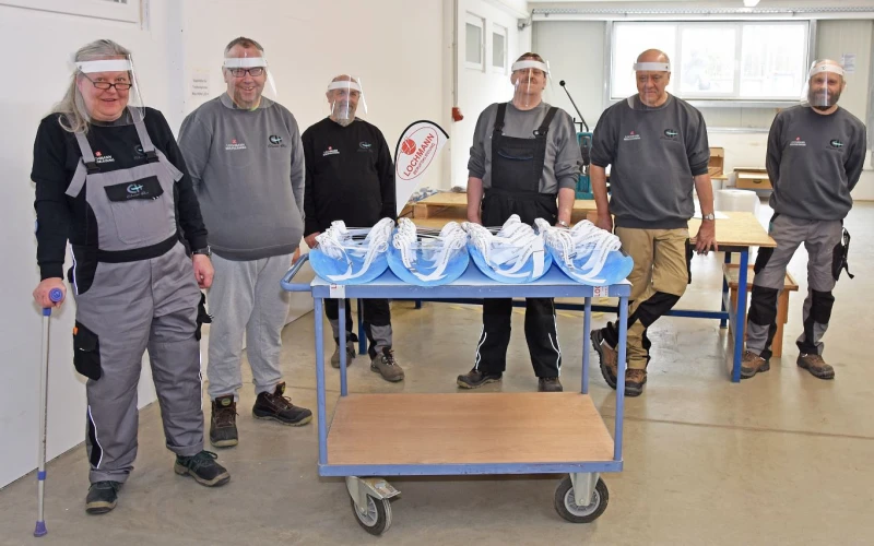 Sechs Personen in Arbeitskleidung mit Gesichtsvisieren stehen hinter einem Transportwagen mit blauen Gesichtsvisieren. Im Hintergrund sind Holztische zu sehen.