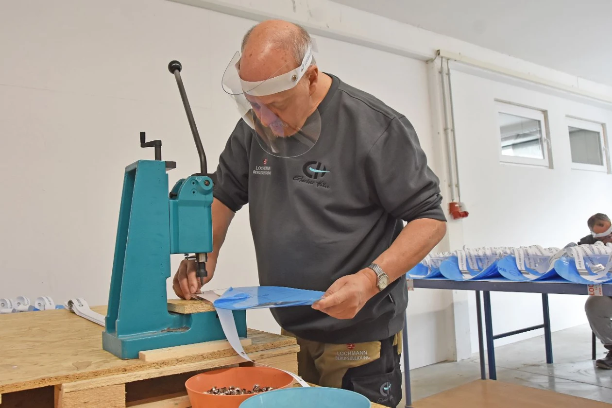 Ein Mann mit Gesichtsvisier und grauem Pullover arbeitet an einer t&uuml;rkisfarbenen Maschine an einem Holztisch. Im Hintergrund sind bereits fertig gestellte Gesichtsvisiere auf einem Tisch drapiert.