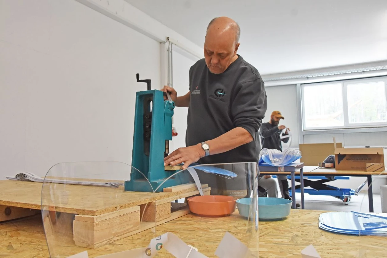 Ein &auml;terer Mann bedient eine t&uuml;rkisfarbene Maschine an einem Holztisch in einer Werkstatt. Er tr&auml;gt ein graues Sweatshirt und konzentriert sich auf seine Arbeit. Im Hintergrund ist eine weitere Person unscharf zu sehen.