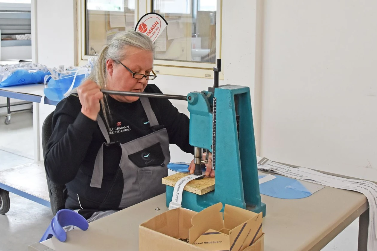 Eine &auml;ltere Frau mit Brille sitzt an einem Arbeitstisch in einer Werkstatt. Sie tr&auml;gt eine graue Arbeitshose und einen schwarzen Pullover. Sie bedient eine blaue Stanzmaschine.