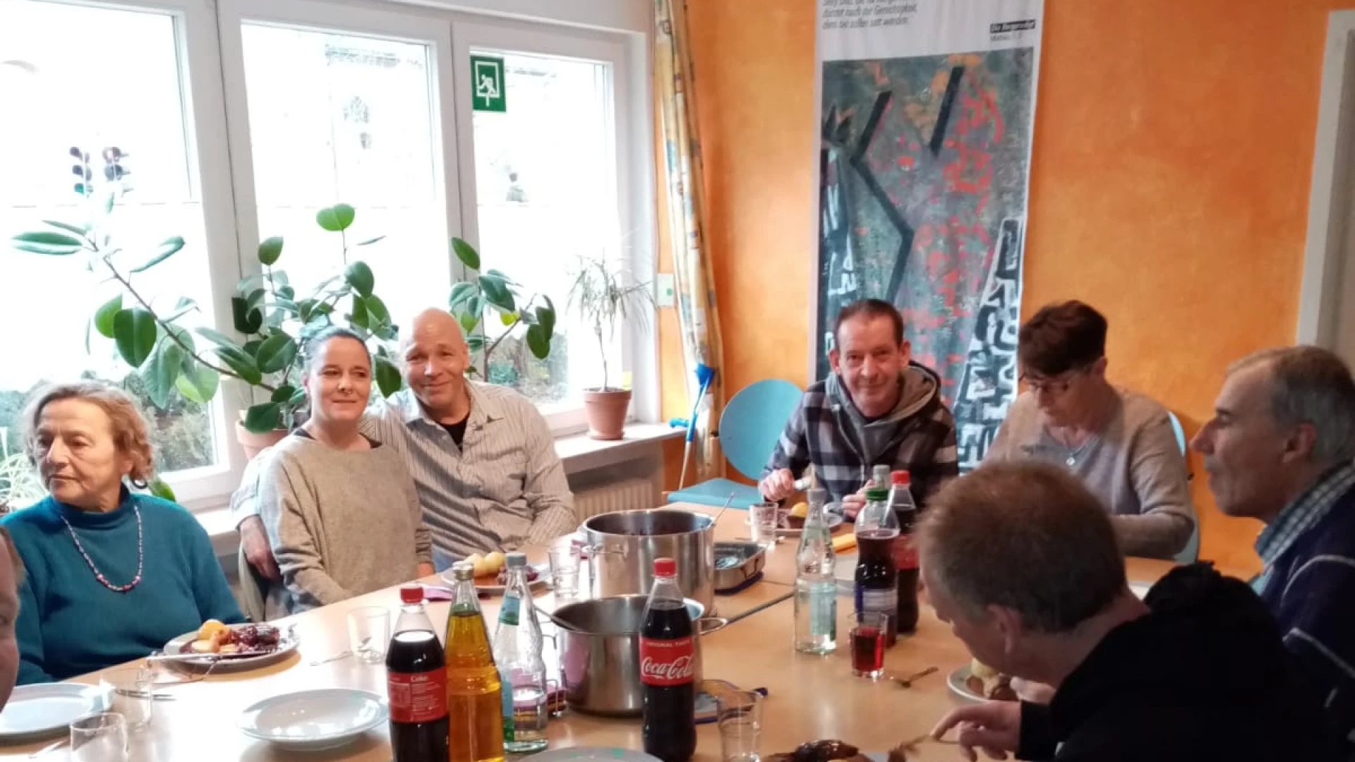 Mehrere Personen sitzen an einem gedeckten Tisch in einem Raum mit einer orangefarbener Wand. Am gro&szlig;en Fenster stehen Pflanzen.