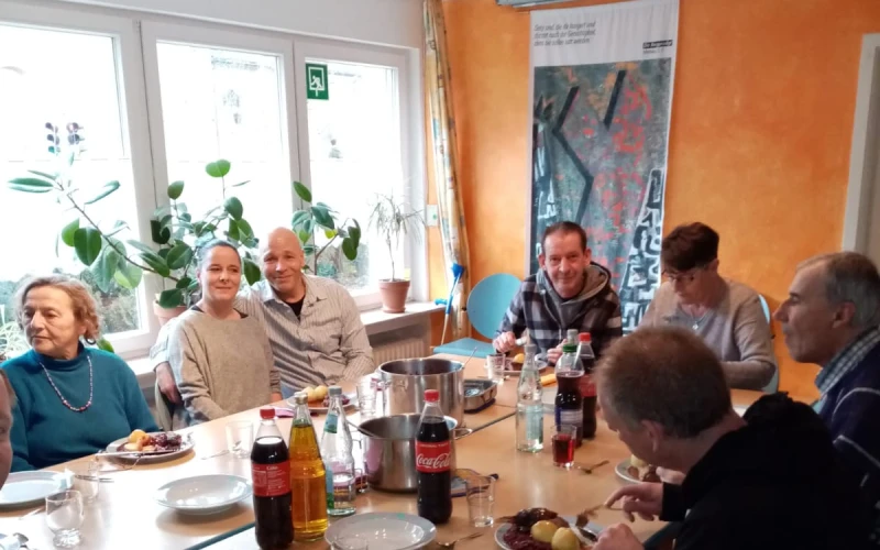 Mehrere Personen sitzen an einem gedeckten Tisch in einem Raum mit einer orangefarbener Wand. Am gro&szlig;en Fenster stehen Pflanzen.