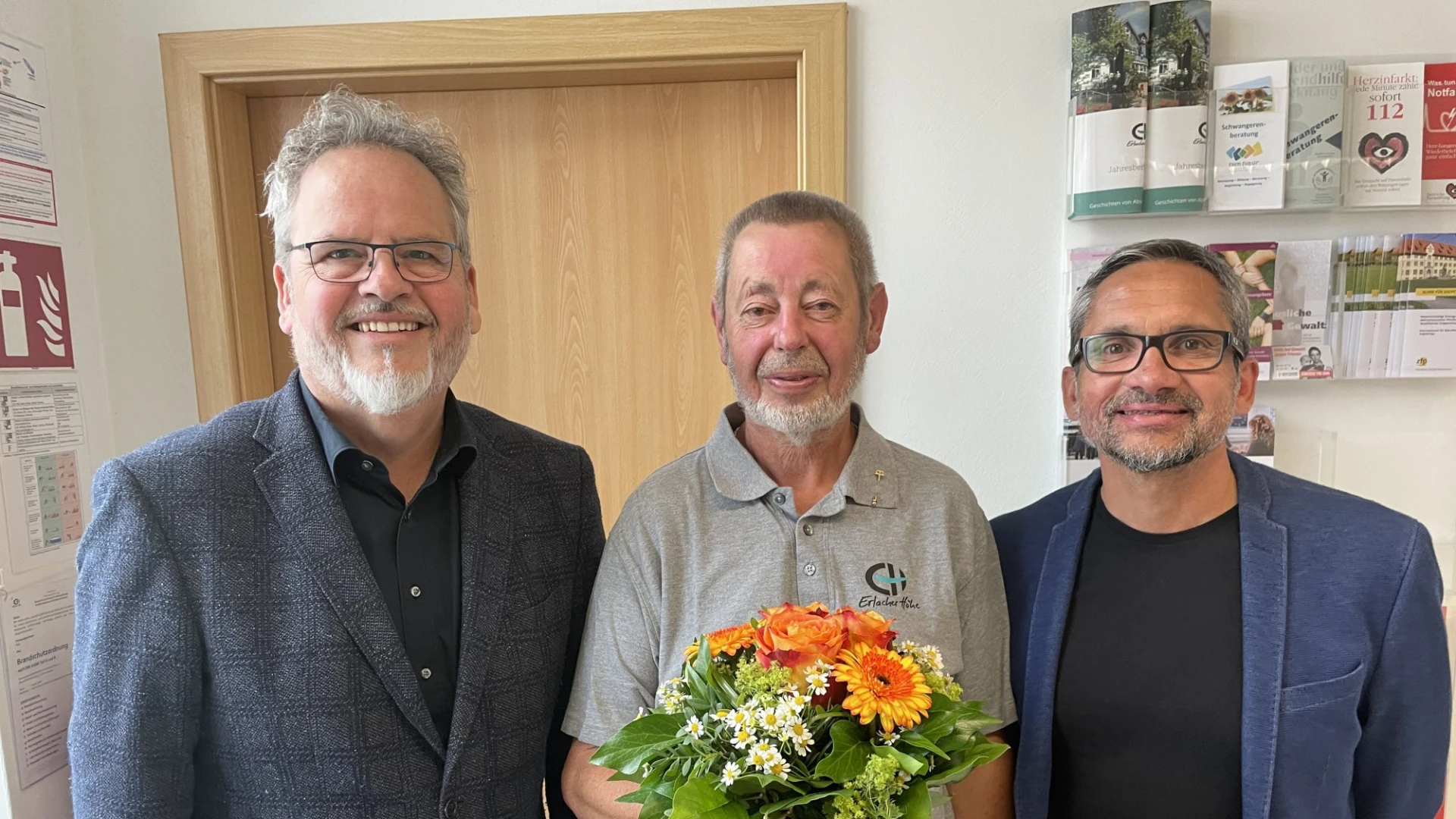 Drei M&auml;nner stehen l&auml;chelnd in die Kamera blickend im Eingangsbereich einer sozialen Einrichtung. Der mittlere &auml;ltere Mann h&auml;lt einen Blumenstrau&szlig; in der Hand. Es scheint sich um ein Dienstjubil&auml;um zu handeln.