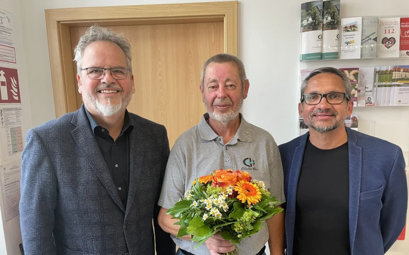 Drei M&auml;nner stehen l&auml;chelnd in die Kamera blickend im Eingangsbereich einer sozialen Einrichtung. Der mittlere &auml;ltere Mann h&auml;lt einen Blumenstrau&szlig; in der Hand. Es scheint sich um ein Dienstjubil&auml;um zu handeln.