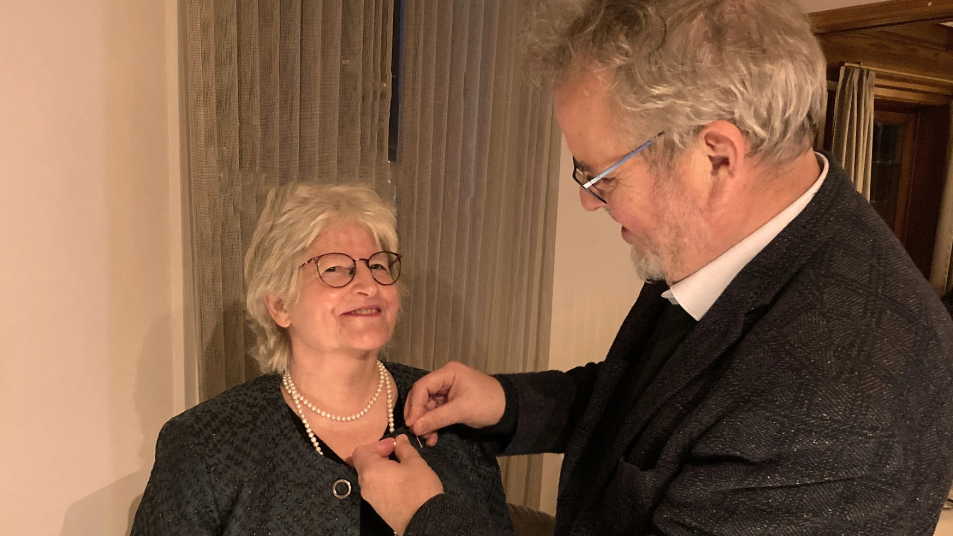 Eine Frau und ein Mann, beide in festlicher Kleidung stehen sich gegen&uuml;ber. Der Mann steckt der Frau, die l&auml;chelnd in die Kamera blickt, das Kronenkreuz ans Rever ihrer Jacke. Beide tragen Brillen und wirken sehr freudig.