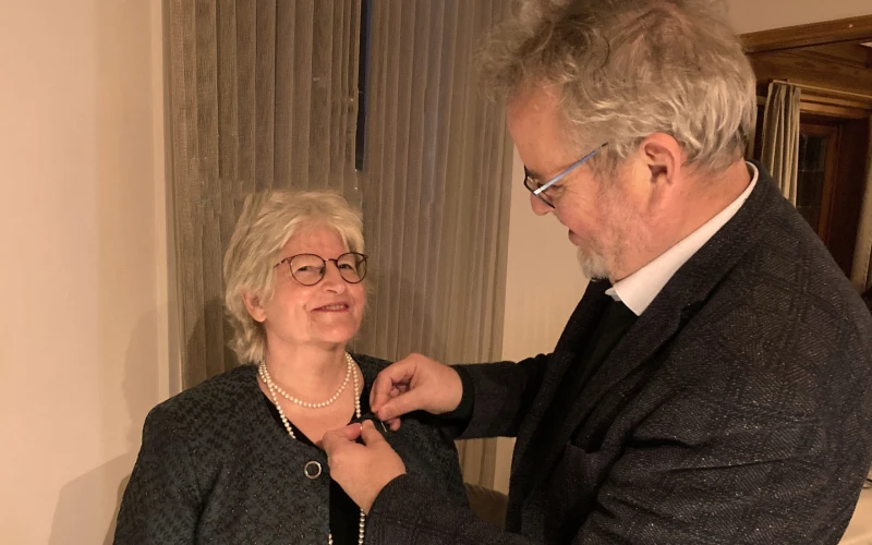 Eine Frau und ein Mann, beide in festlicher Kleidung stehen sich gegen&uuml;ber. Der Mann steckt der Frau, die l&auml;chelnd in die Kamera blickt, das Kronenkreuz ans Rever ihrer Jacke. Beide tragen Brillen und wirken sehr freudig.