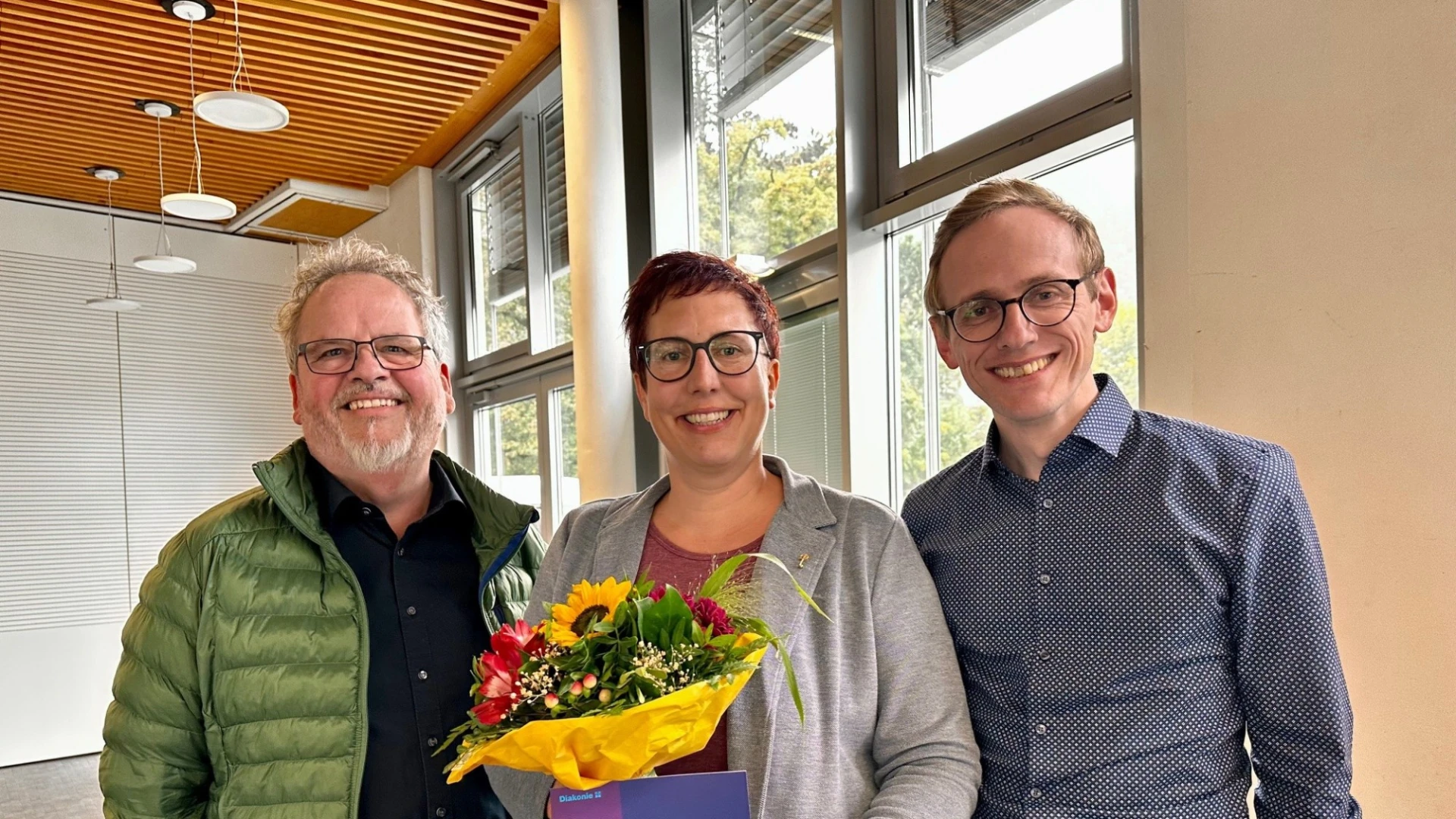 Eine Frau erh&auml;lt das Goldene Kronenkreuz und wird von zwei M&auml;nnern bei der &Uuml;bergabe gratuliert, w&auml;hrend sie einen Blumenstrau&szlig; und eine Urkunde in den H&auml;nden h&auml;lt.