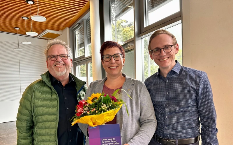 Eine Frau erh&auml;lt das Goldene Kronenkreuz und wird von zwei M&auml;nnern bei der &Uuml;bergabe gratuliert, w&auml;hrend sie einen Blumenstrau&szlig; und eine Urkunde in den H&auml;nden h&auml;lt.