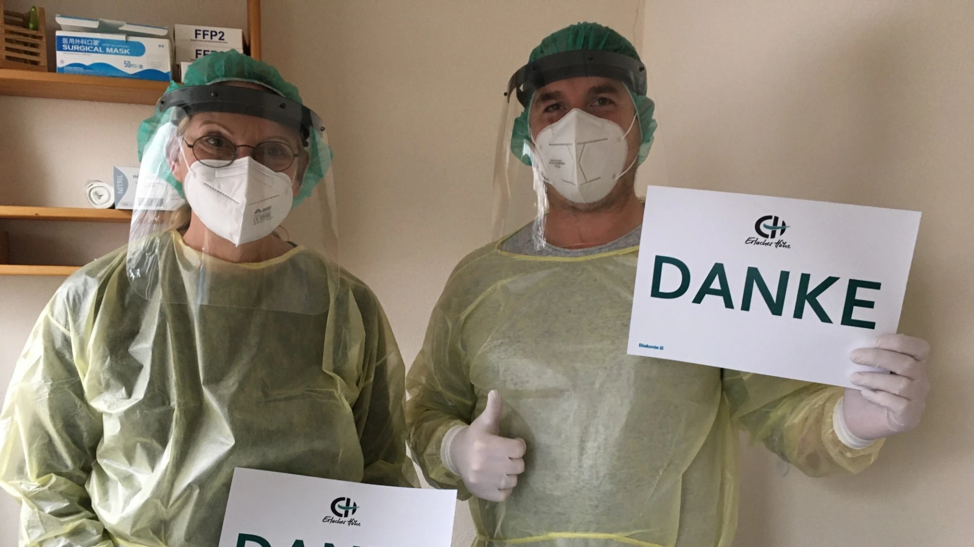 Zwei Personen in vollst&auml;ndiger Schutzkleidung mit Gesichtsvisier, Mund-Nasenschutz und Handschuhe halten jeweils ein Blatt Papier mit der Aufschrift "Danke" in den H&auml;nden.
