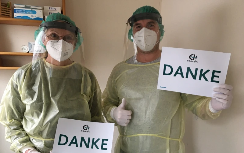 Zwei Personen in vollst&auml;ndiger Schutzkleidung mit Gesichtsvisier, Mund-Nasenschutz und Handschuhe halten jeweils ein Blatt Papier mit der Aufschrift "Danke" in den H&auml;nden.