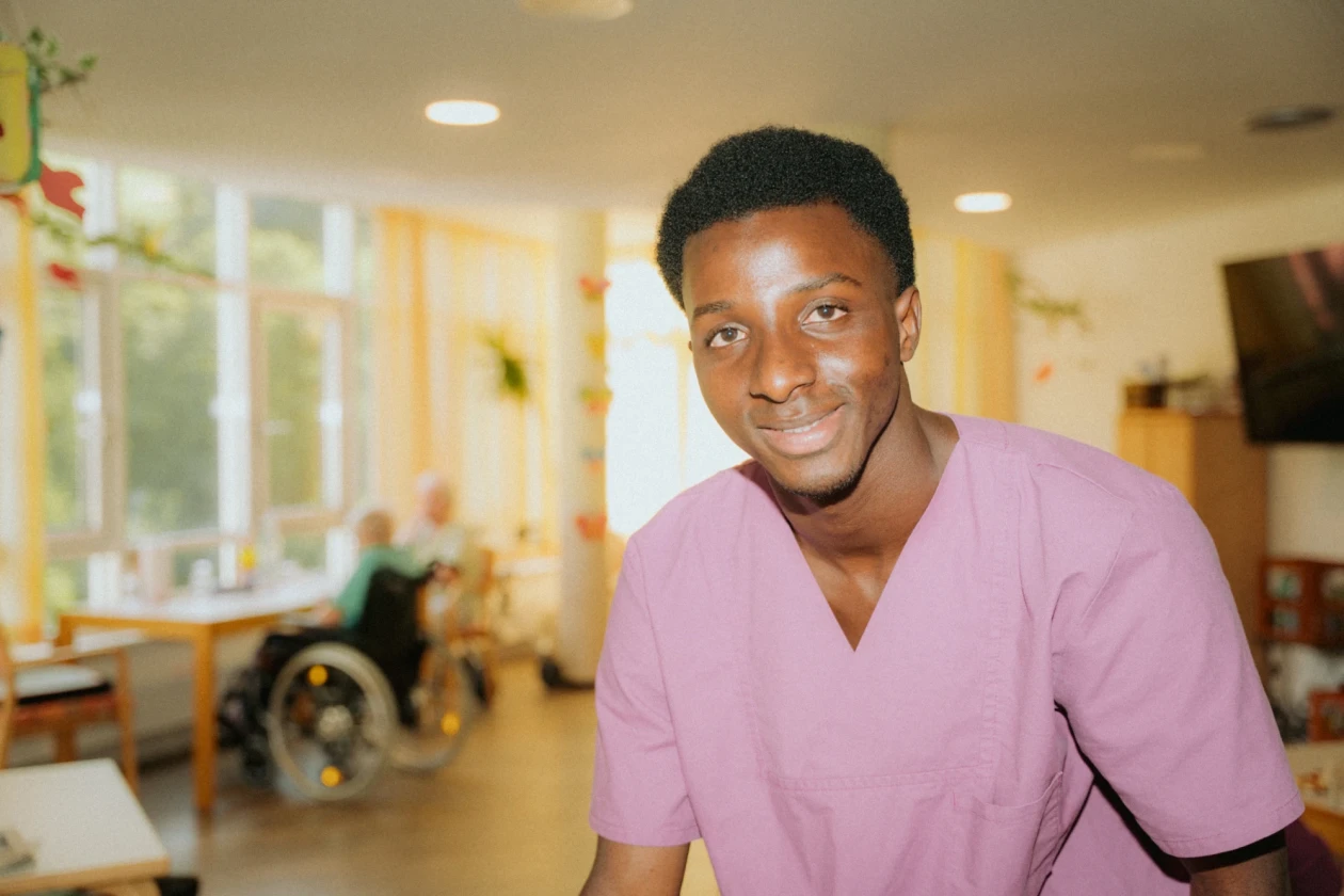 Ein l&auml;chelnder junger Mann in pinkem medizinischem Kittel steht in einer hellen Einrichtung mit Behandlungszimmern im Hintergrund.