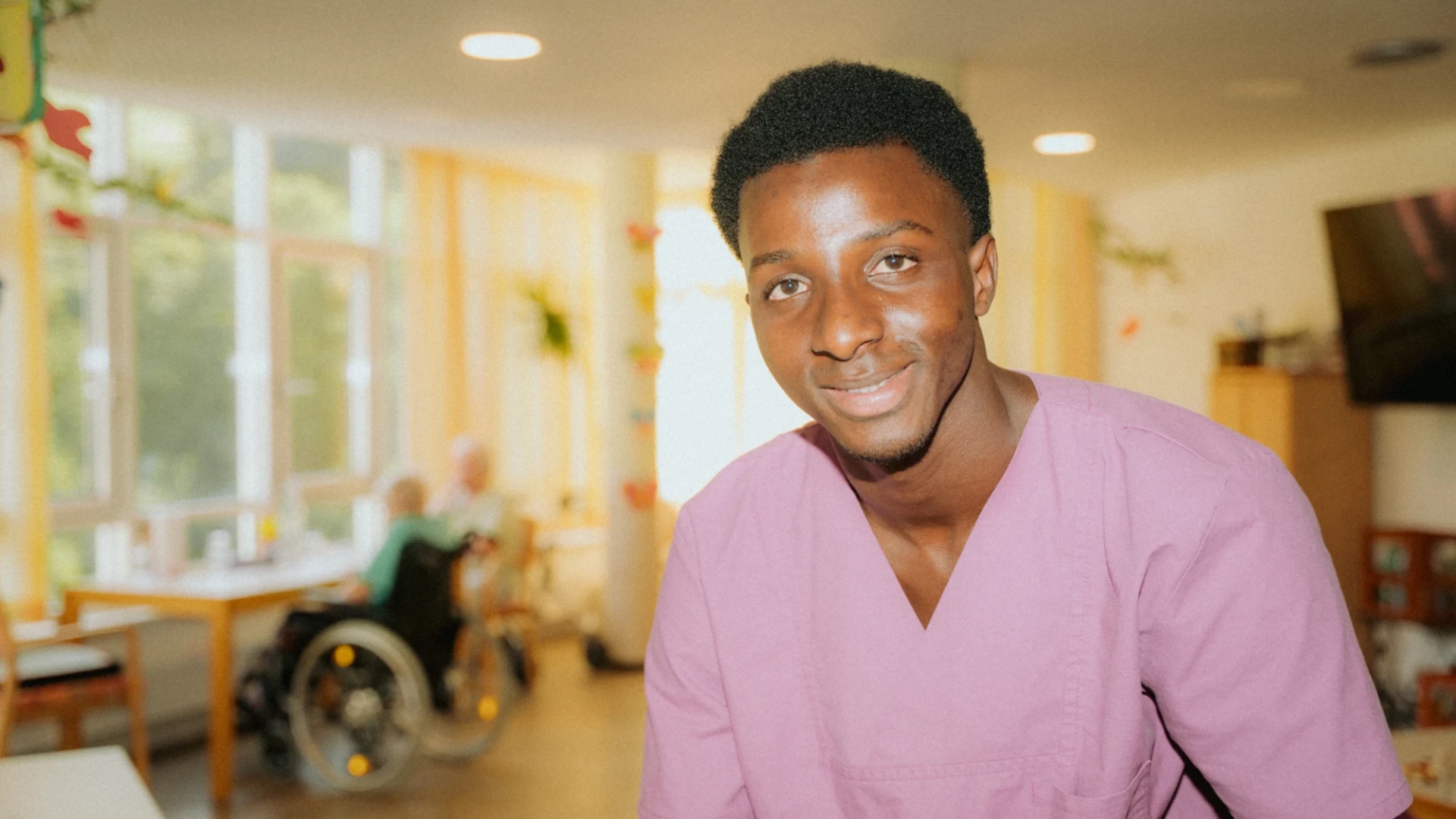 Ein l&auml;chelnder junger Mann in pinkem medizinischem Kittel steht in einer hellen Einrichtung mit Behandlungszimmern im Hintergrund.