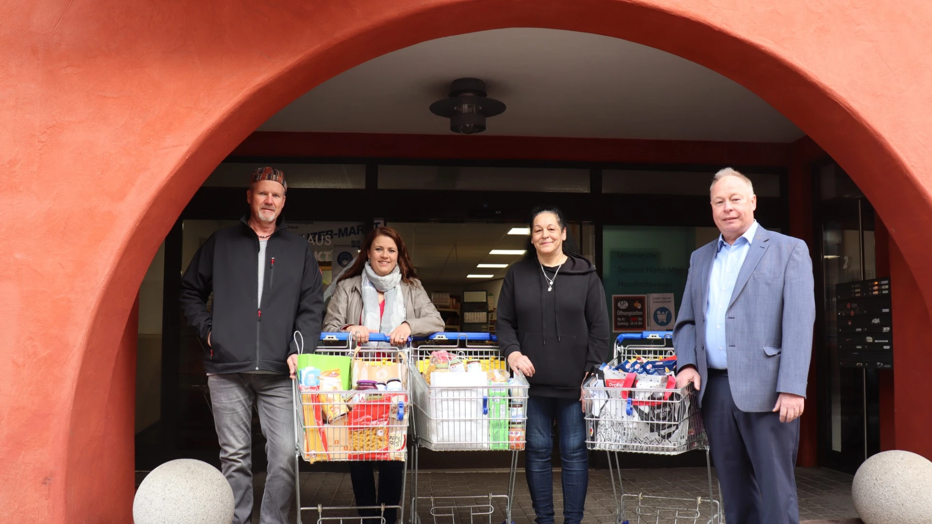 Vier Personen stehen unter einem roten Torbogen. Vor ihnen stehen drei Einkaufswagen voller Lebensmittel.