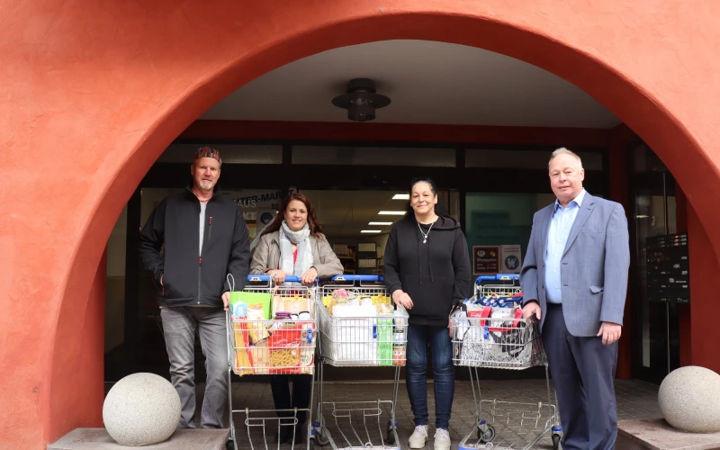 Vier Personen stehen unter einem roten Torbogen. Vor ihnen stehen drei Einkaufswagen voller Lebensmittel.