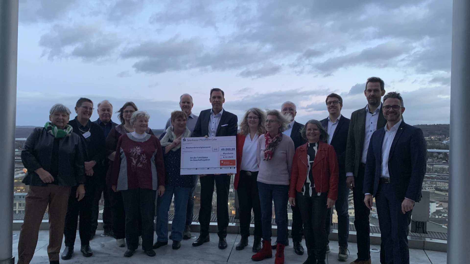 Eine Menschengruppe steht auf einer Terrasse. In der Mitte ist ein Spendenscheck zu sehen, der &uuml;bergeben wurde. Im Hintergrund ist die Kulisse einer Stadt mit einem sehr bew&ouml;lkten Himmel zu sehen.