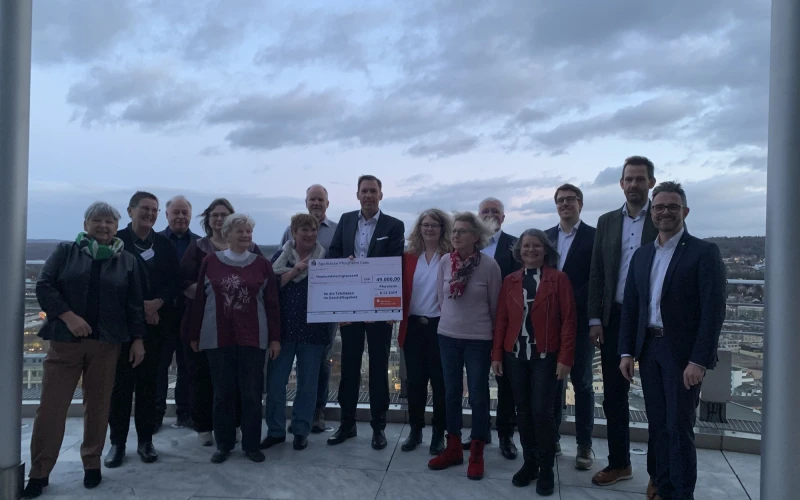 Eine Menschengruppe steht auf einer Terrasse. In der Mitte ist ein Spendenscheck zu sehen, der &uuml;bergeben wurde. Im Hintergrund ist die Kulisse einer Stadt mit einem sehr bew&ouml;lkten Himmel zu sehen.