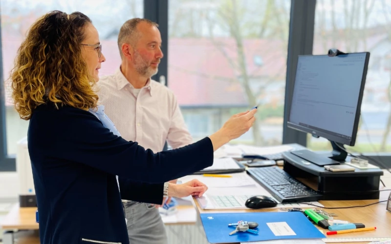 Zwei Personen in einem B&uuml;roraum mit gro&szlig;en Fenstern, die gemeinsam an einem Computer arbeiten. Die Frau im dunklen Blazer steht und zeigt auf den Bildschirm, w&auml;hrend der Mann in wei&szlig;em Hemd daneben steht.
