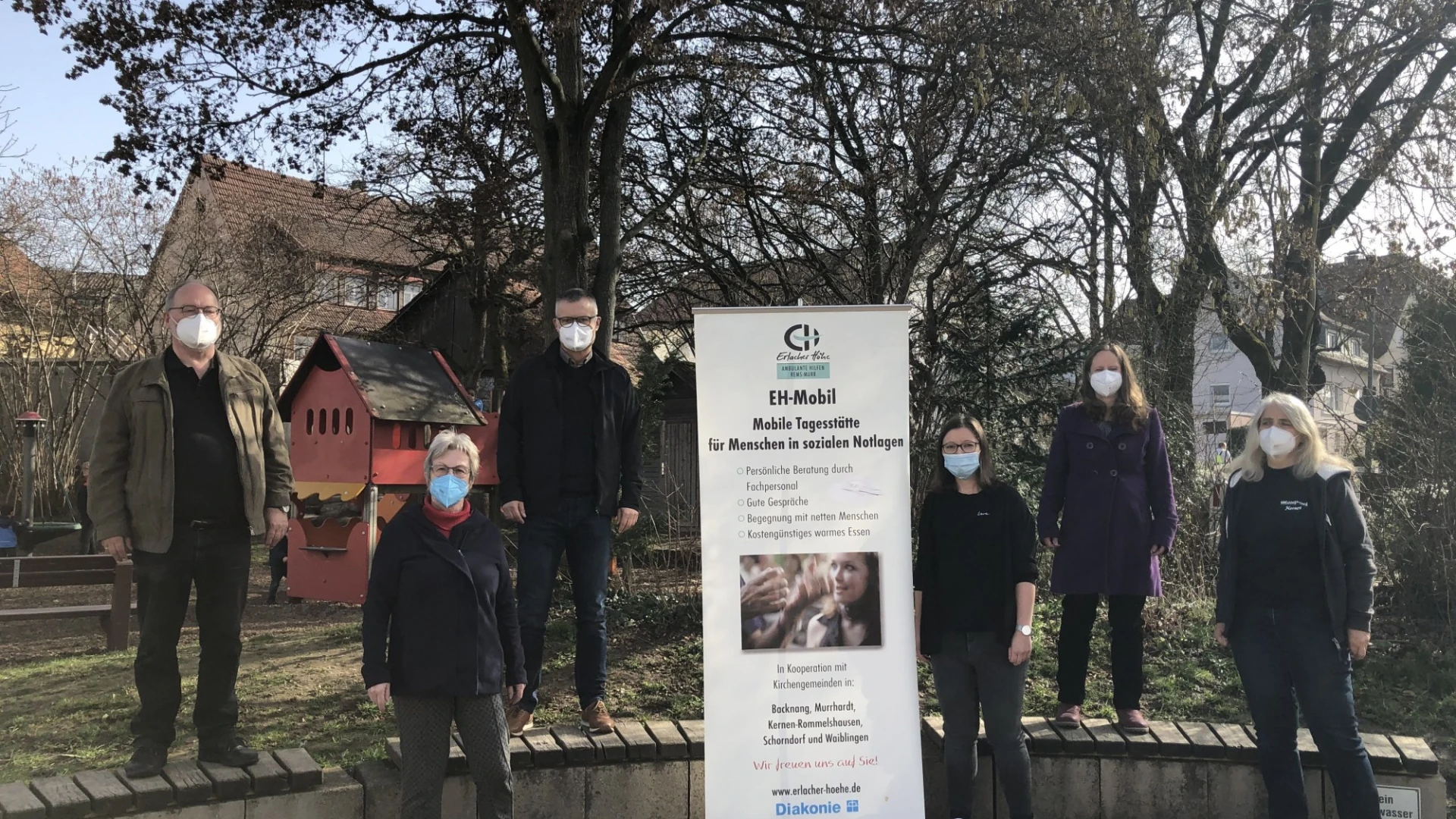 Sechs Personen mit Mund-Nasenschutz stehen im Freien um ein Banner mit Informationen zum "EH Mobil". Hinter ihnen sind B&auml;ume und H&auml;user zu sehen.