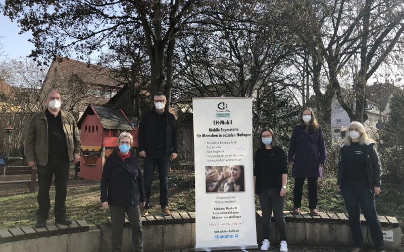 Sechs Personen mit Mund-Nasenschutz stehen im Freien um ein Banner mit Informationen zum "EH Mobil". Hinter ihnen sind B&auml;ume und H&auml;user zu sehen.