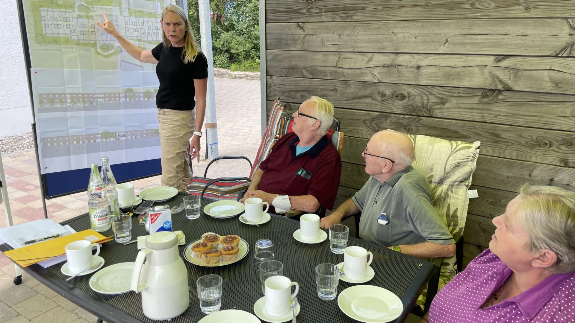 Frau mit blonden Haaren pr&auml;sentiert an einem Whiteboard, w&auml;hrend drei &auml;ltere Menschen an einem gedeckten Tisch sitzen und ihr zuh&ouml;ren, offenbar in einer Besprechung im Freien.
