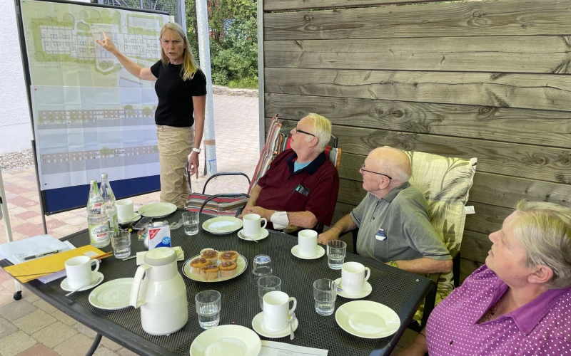 Frau mit blonden Haaren pr&auml;sentiert an einem Whiteboard, w&auml;hrend drei &auml;ltere Menschen an einem gedeckten Tisch sitzen und ihr zuh&ouml;ren, offenbar in einer Besprechung im Freien.