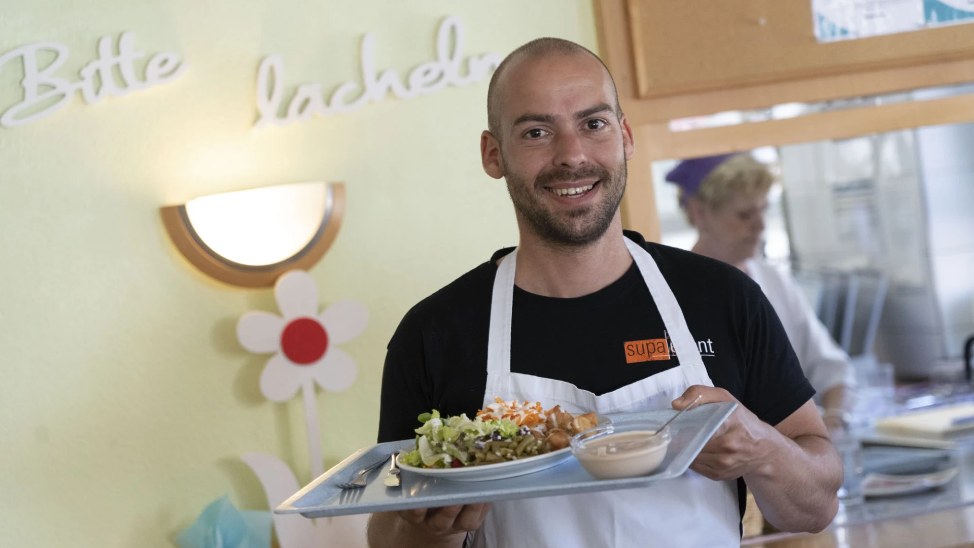 Mann in wei&szlig;er Sch&uuml;rze l&auml;chelt und h&auml;lt ein Tablett mit Speisen, im Hintergrunddekorierte Wand mi Lampe und eine Frau in wei&szlig;er Kleidung in einer K&uuml;che.