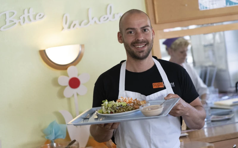 Mann in wei&szlig;er Sch&uuml;rze l&auml;chelt und h&auml;lt ein Tablett mit Speisen, im Hintergrunddekorierte Wand mi Lampe und eine Frau in wei&szlig;er Kleidung in einer K&uuml;che.