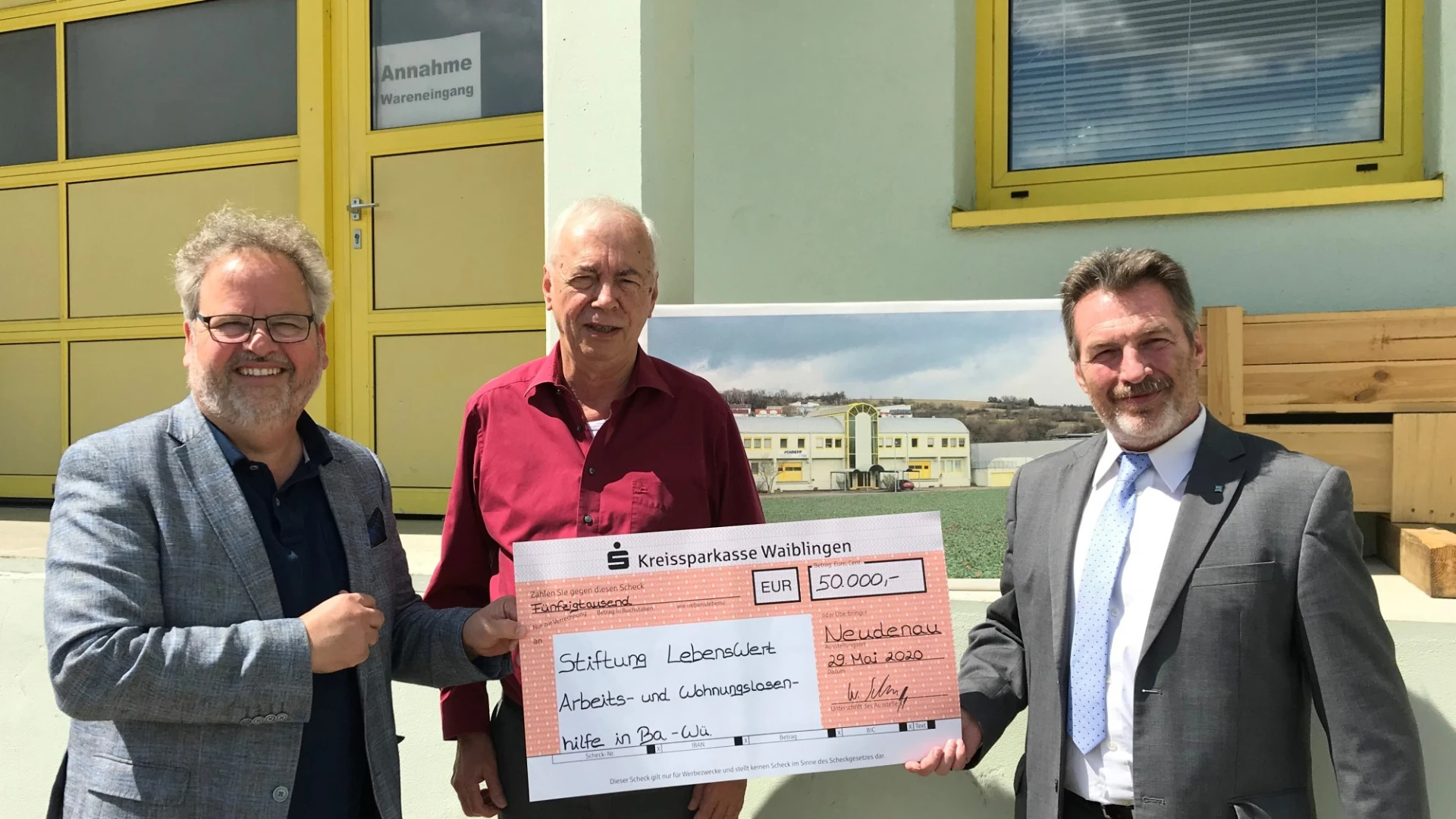 Drei M&auml;nner halten einen Scheck in der Hand und lachen in die Kamera. Im Hintergrund ist ein Fabrikgeb&auml;ude mit gelben Fensterrahmen und Eingangstor zu sehen.