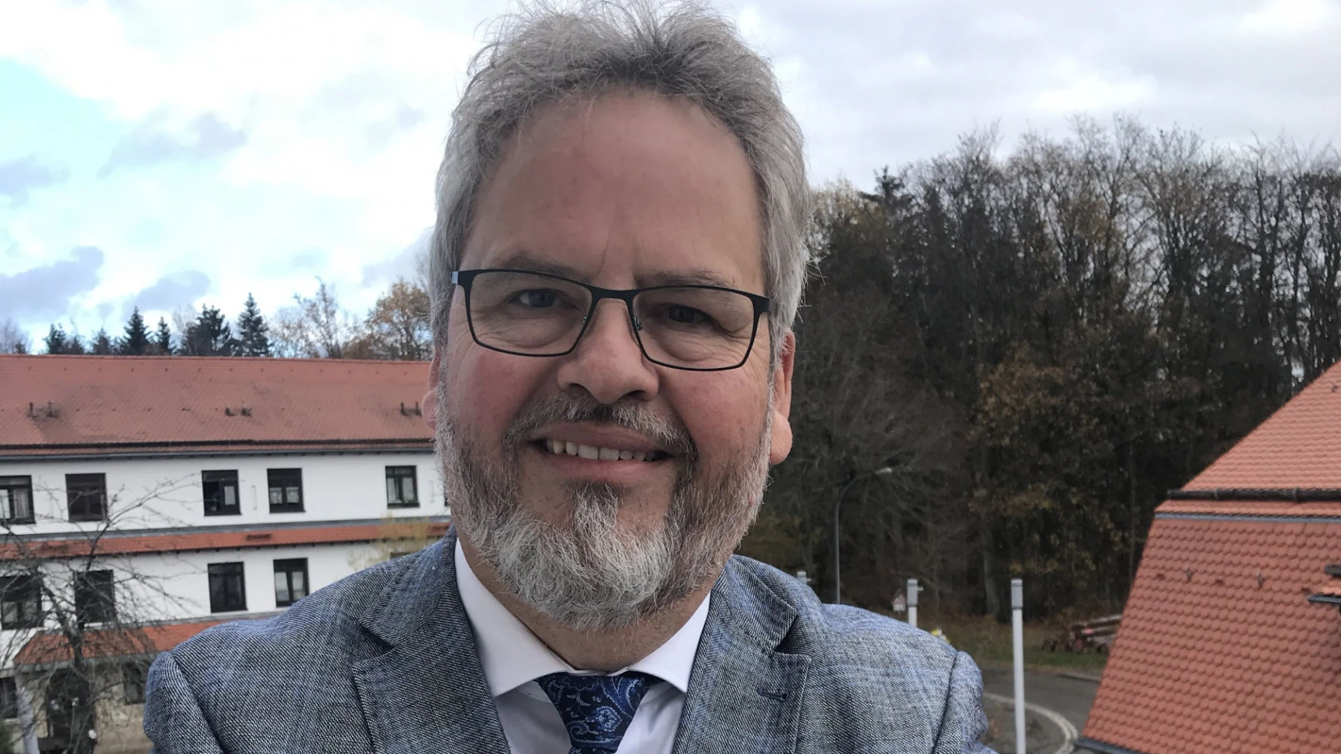 Ein &auml;lterer Mann mit grauem Anzug, blauer Krawatte, wei&szlig;em Hemd und Brille steht auf einem Balkon. Im Hintergrund sind zwei weitere Geb&auml;ude und eine Stra&szlig;e zu sehen.