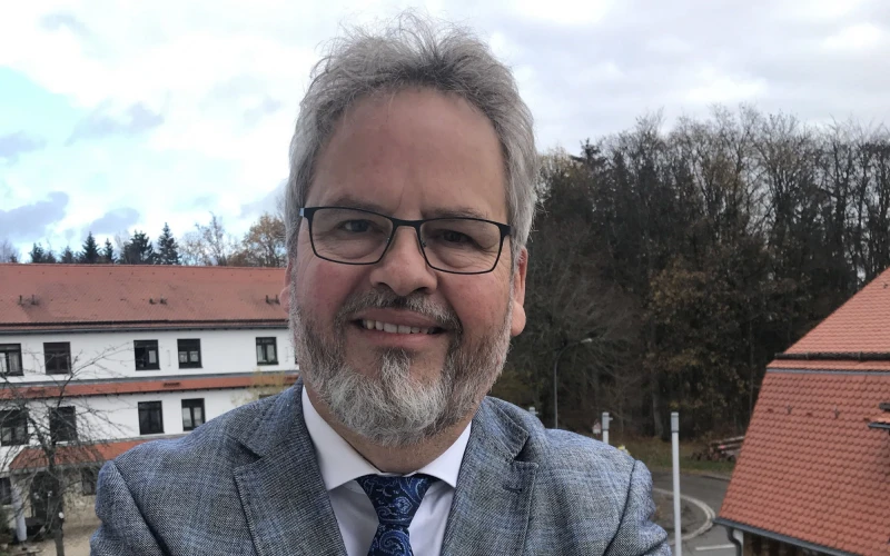 Ein &auml;lterer Mann mit grauem Anzug, blauer Krawatte, wei&szlig;em Hemd und Brille steht auf einem Balkon. Im Hintergrund sind zwei weitere Geb&auml;ude und eine Stra&szlig;e zu sehen.