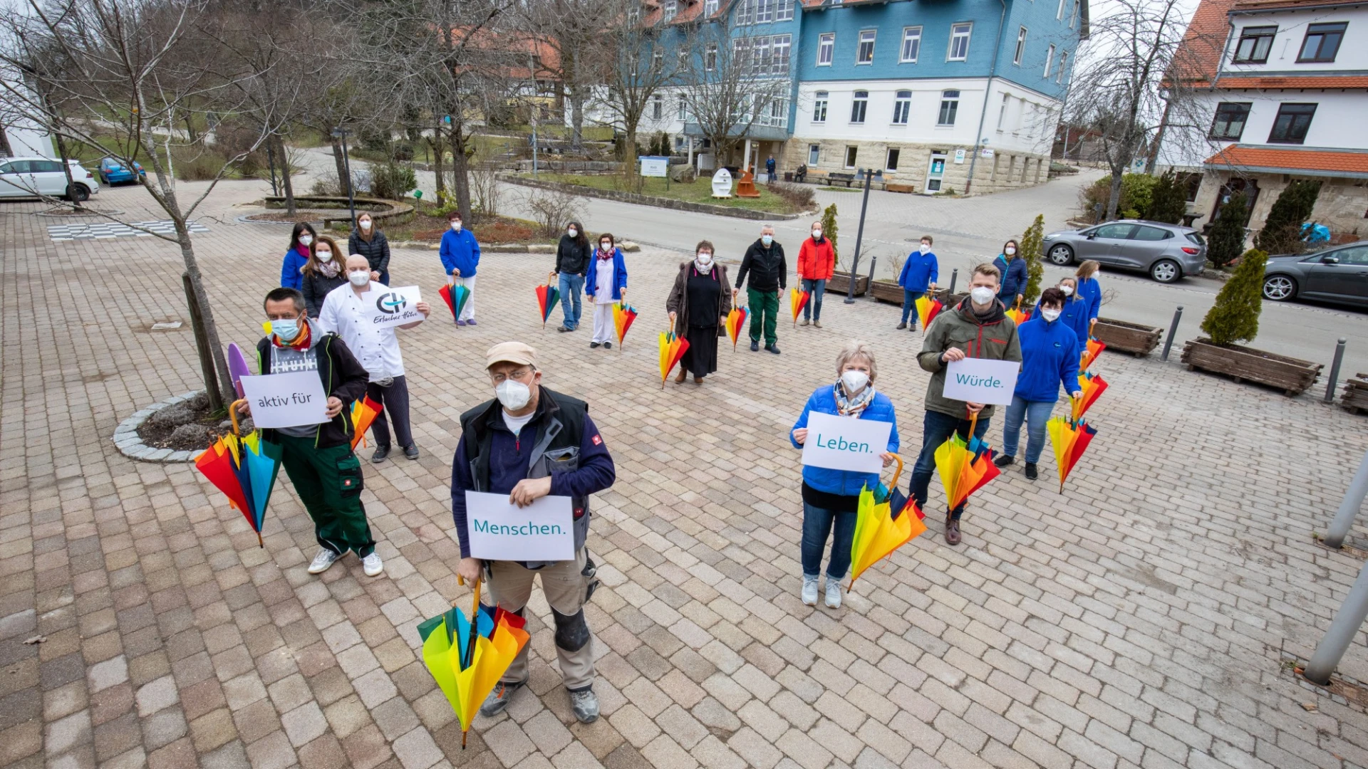 Mehrere Personen stehen auf einer gepflasterten Hoffl&auml;che und halten bunte Regenschirme sowie Schilder mit den W&ouml;rtern "aktiv f&uuml;r", "Menschen", "Leben", und "W&uuml;rde". Alle tragen medizinische Masken und im Hintergrund sind mehrere Geb&auml;ude und B&auml;ume zu sehen.