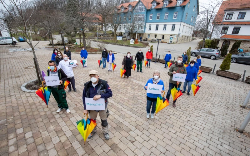 Mehrere Personen stehen auf einer gepflasterten Hoffl&auml;che und halten bunte Regenschirme sowie Schilder mit den W&ouml;rtern "aktiv f&uuml;r", "Menschen", "Leben", und "W&uuml;rde". Alle tragen medizinische Masken und im Hintergrund sind mehrere Geb&auml;ude und B&auml;ume zu sehen.