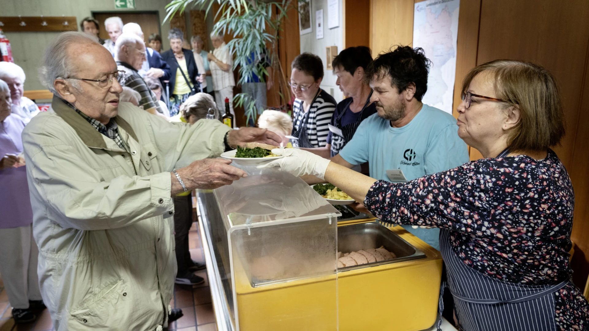 Ein &auml;lterer Mann in beiger Jacke bekommt von einer Frau in dunklem Blazer &uuml;ber einen gelben Beh&auml;lter hinweg einen Teller mit Essen gereicht