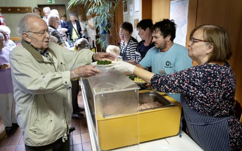 Ein &auml;lterer Mann in beiger Jacke bekommt von einer Frau in dunklem Blazer &uuml;ber einen gelben Beh&auml;lter hinweg einen Teller mit Essen gereicht