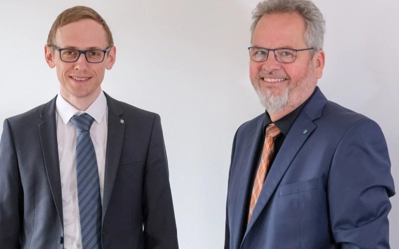 Zwei M&auml;nner mit Brillen in Anz&uuml;gen und Krawatte l&auml;cheln in die Kamera.