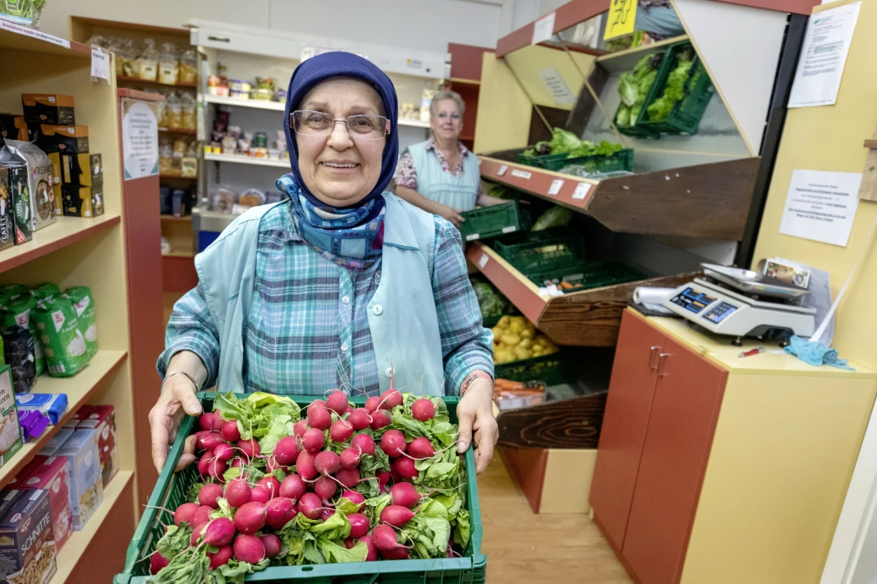 Frau mit Kopftuch und Brille in blau-karierter Bluse und hellblauer Weste pr&auml;sentiert eine Kiste voller roter Radieschen. Im Hintergrund sind Regale gef&uuml;llt mit Lebensmittel zu sehen, sowie eine weitere Frau an einem Gem&uuml;seregal.