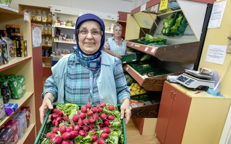 Frau mit Kopftuch und Brille in blau-karierter Bluse und hellblauer Weste pr&auml;sentiert eine Kiste voller roter Radieschen. Im Hintergrund sind Regale gef&uuml;llt mit Lebensmittel zu sehen, sowie eine weitere Frau an einem Gem&uuml;seregal.