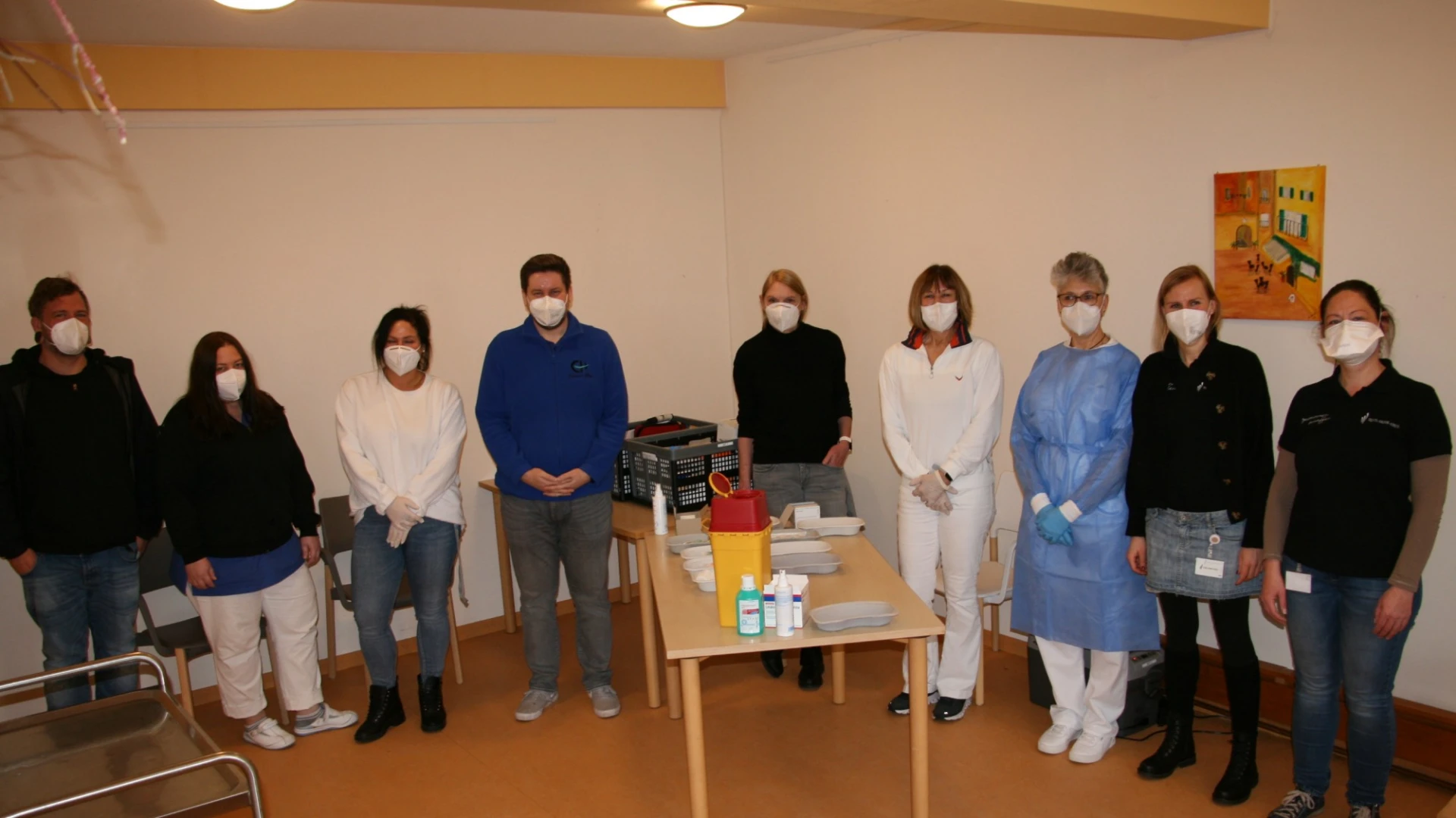 Neun Personen stehen in einem Raum in einer Reihe hinter einem Tisch mit medizinischem Material. Alle tragen Mund-Nasenschutz.