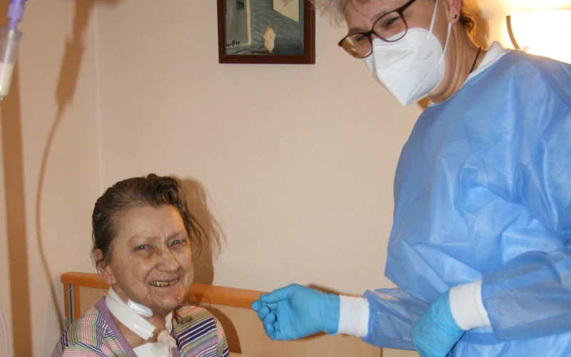 Eine Pflegekraft in blauer Schutzkleidung, Handschuhen und Mund-Nasenschutz steht neben einer im Pflegebett sitzenden, l&auml;chelnden Frau in einem Zimmer.