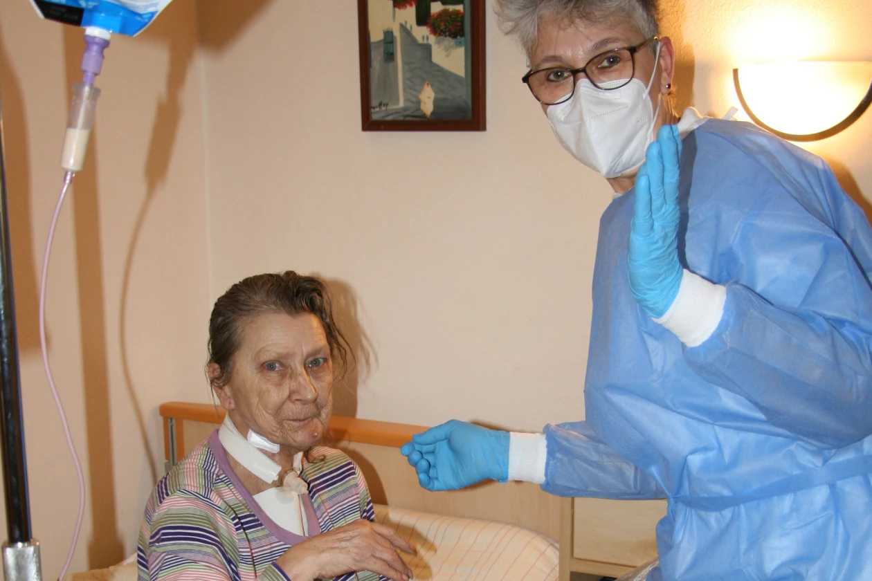 Eine Pflegekraft in blauer Schutzkleidung, Handschuhen und Mund-Nasenschutz steht neben einer im Pflegebett sitzenden Frau in einem Zimmer. Im Hintergrund sind eine Infusion, ein Wandbild und eine Lampe zu sehen.