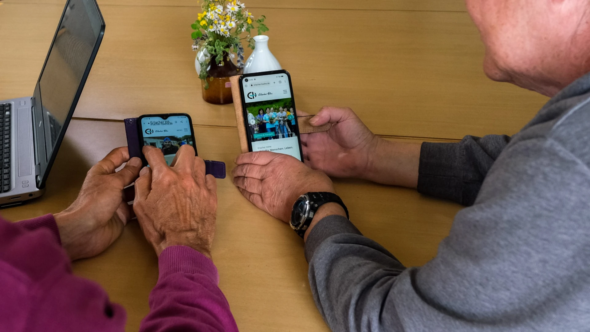 Zwei Personen sitzen an einem Tisch, die Kamera fotografiert &uuml;ber die Schultern. Beide halten ein Handy in der Hand. Auf dem Tisch steht ein Laptop.