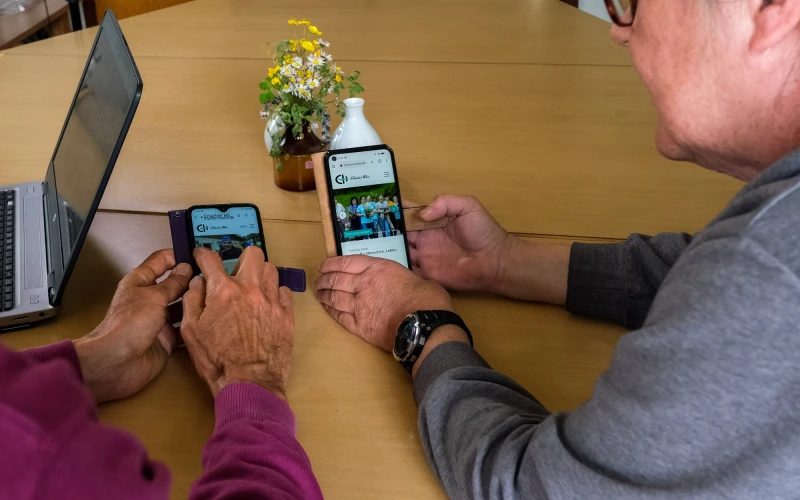 Zwei Personen sitzen an einem Tisch, die Kamera fotografiert &uuml;ber die Schultern. Beide halten ein Handy in der Hand. Auf dem Tisch steht ein Laptop.