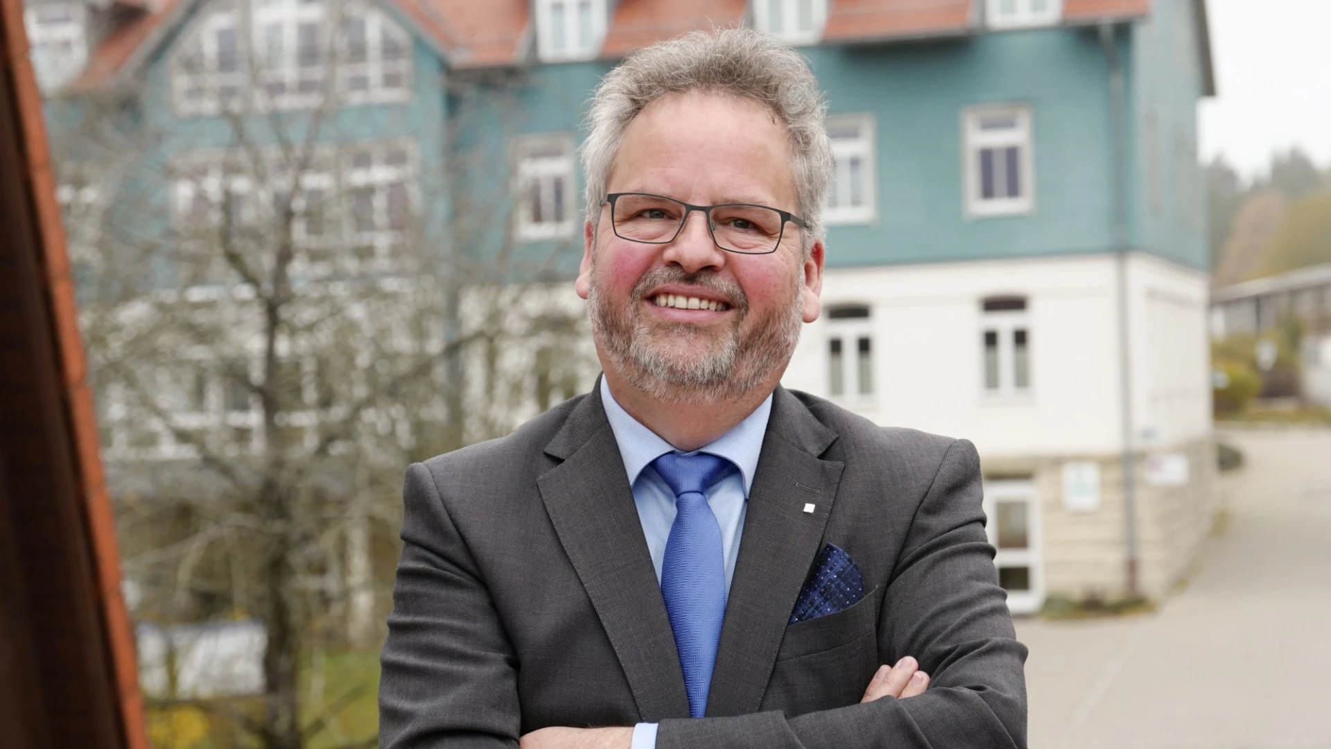 Ein &auml;lterer Mann mit Brille steht mit verschr&auml;nkten Armen vor einem Geb&auml;ude. Er tr&auml;gt einen grauen Anzug, eine blaue Krawatte und ein hellblaues Hemd.