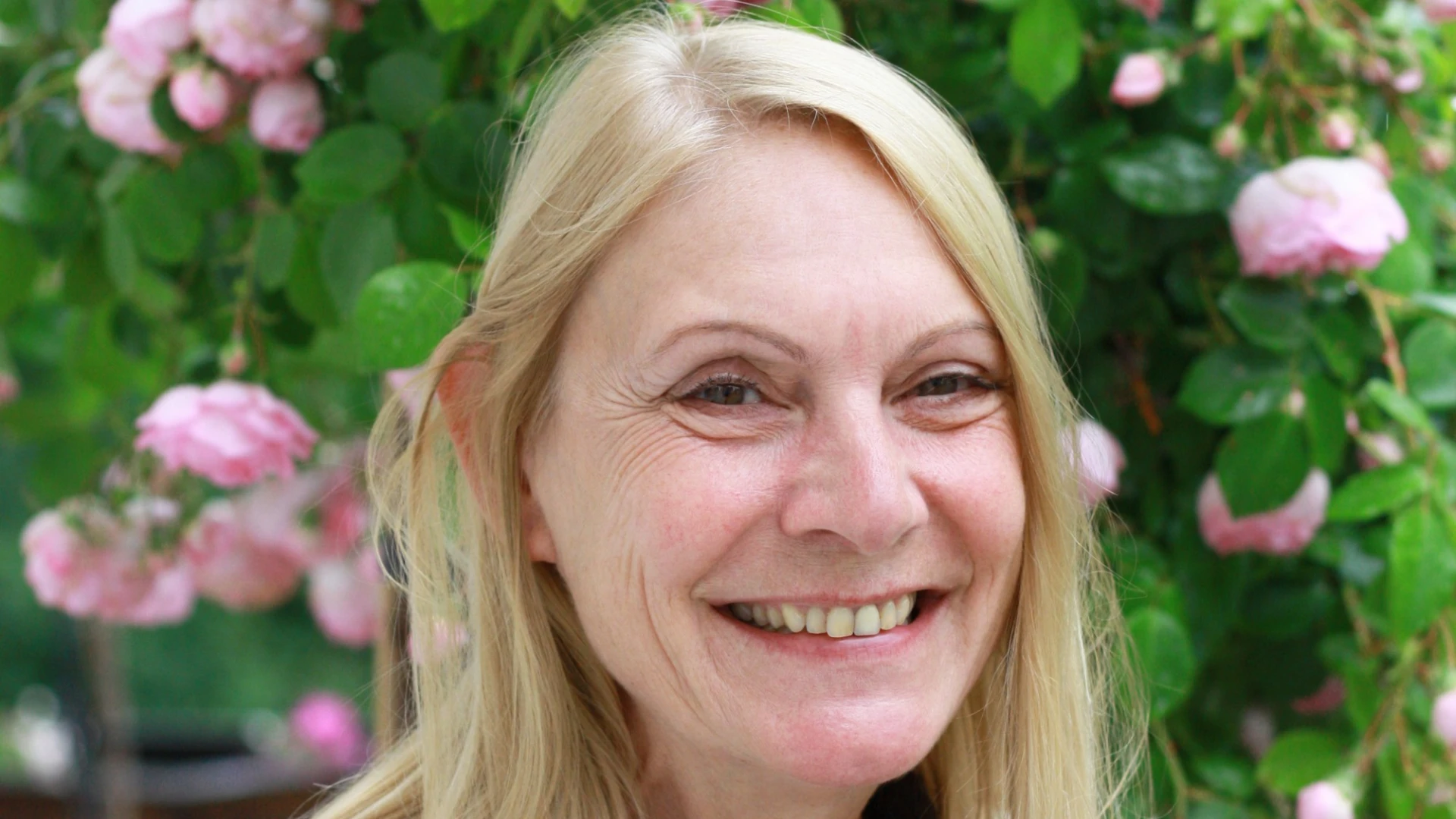 Eine Frau mit blonden glatten Haaren l&auml;chelt in die Kamera. Sie tr&auml;gt ein dunkles Oberteil und dar&uuml;ber eine wei&szlig;e Strickjacke. Im Hintergrund ist ein Rosenstock zu sehen.