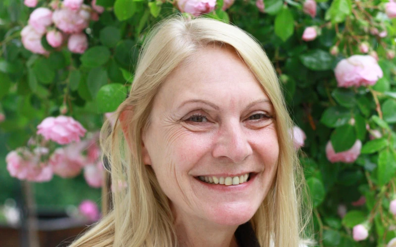 Eine Frau mit blonden glatten Haaren l&auml;chelt in die Kamera. Sie tr&auml;gt ein dunkles Oberteil und dar&uuml;ber eine wei&szlig;e Strickjacke. Im Hintergrund ist ein Rosenstock zu sehen.