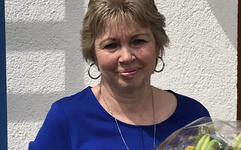 Eine Frau mit kurzen blonden Haaren l&auml;chelt in die Kamera und h&auml;lt einen Blumenstrau&szlig; in der Hand. Sie tr&auml;gt ein blaues Oberteil und eine Halskette.