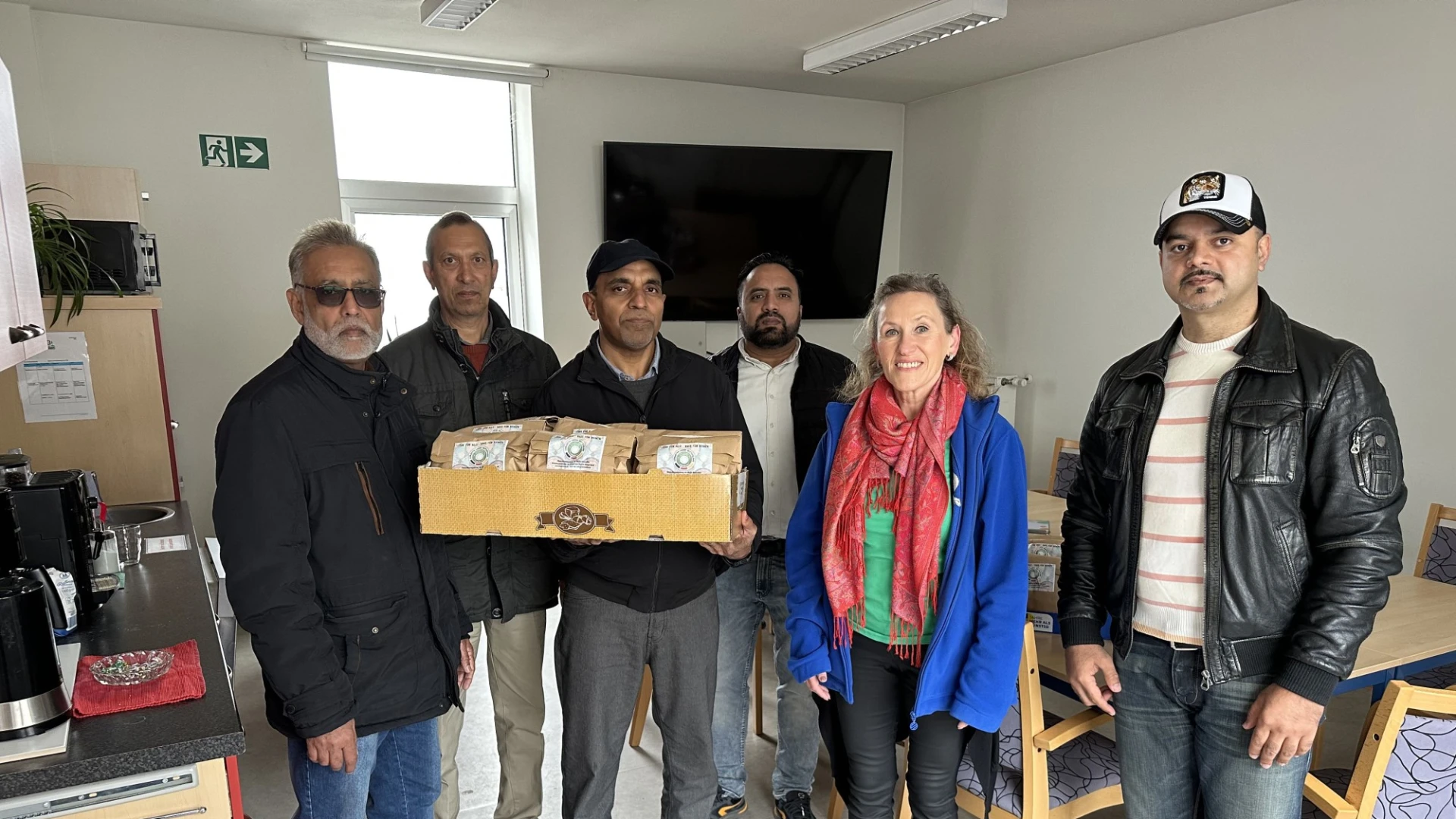 Sechs Personen stehen in einem B&uuml;roraum und halten eine Holzkiste. Sie posieren f&uuml;r ein Gruppenfoto vor einem Fenster und einem Fernseher.