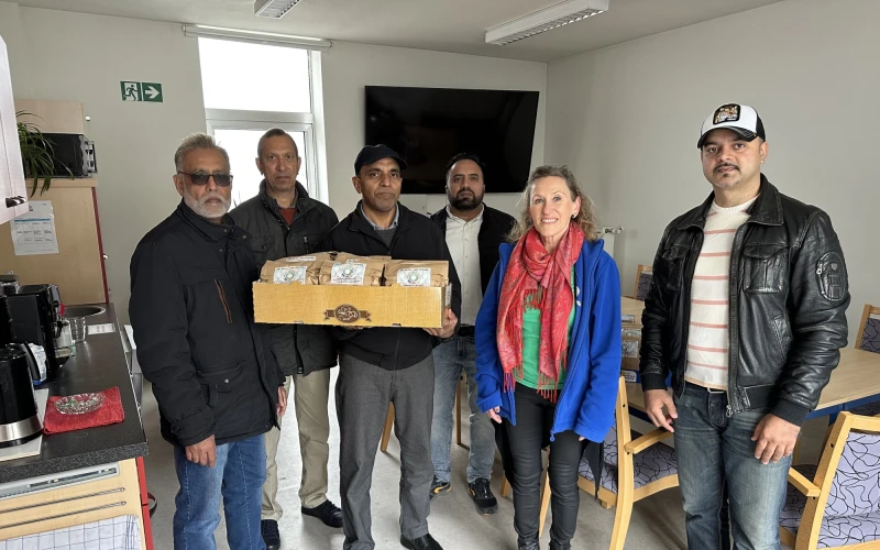 Sechs Personen stehen in einem B&uuml;roraum und halten eine Holzkiste. Sie posieren f&uuml;r ein Gruppenfoto vor einem Fenster und einem Fernseher.
